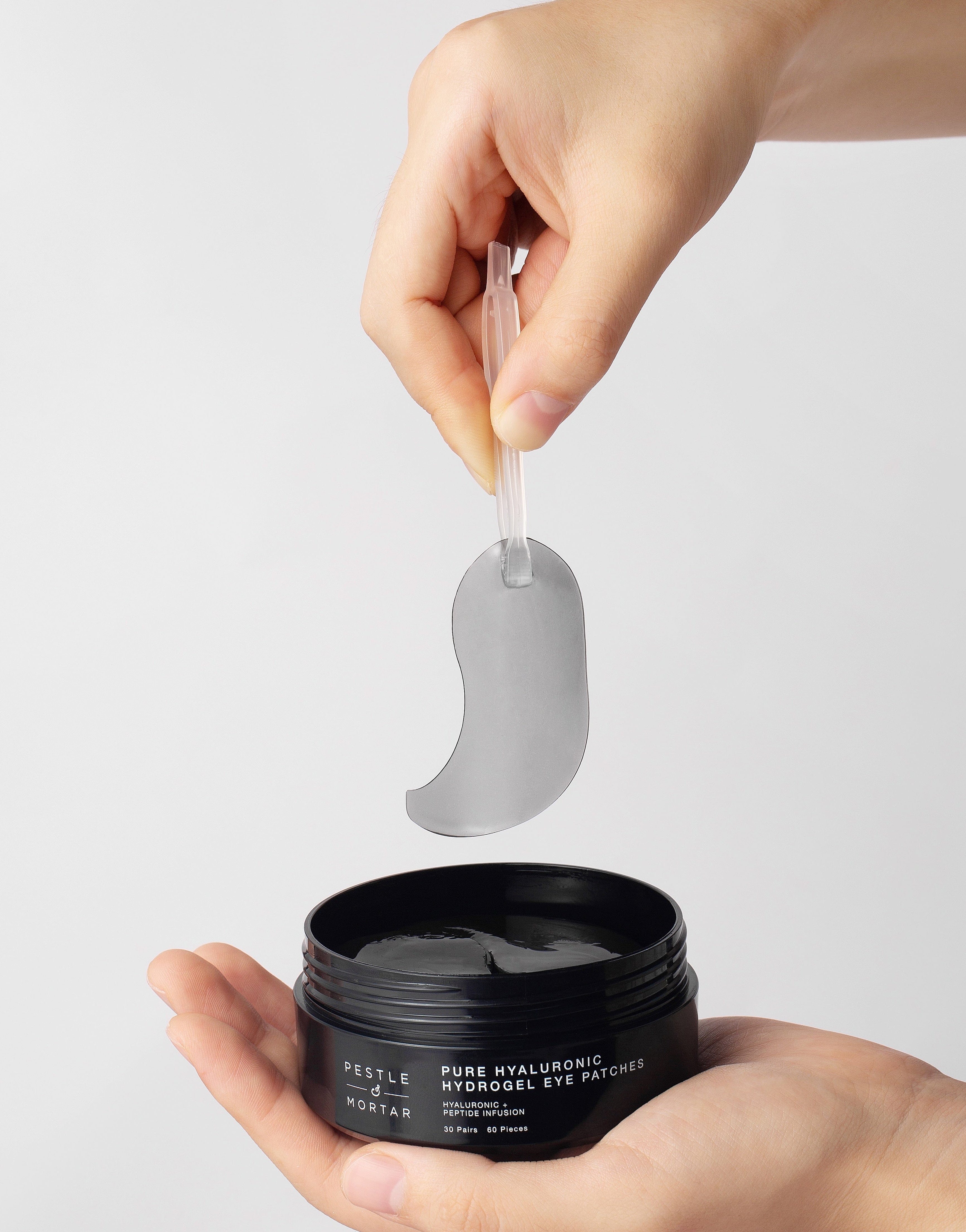Person holding a black jar of Pestle & mortar Pure hyaluronic Hydrogel Eye Patches with a eye patch above it on a light gray background
